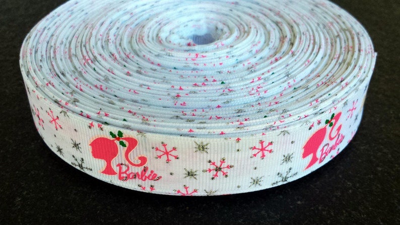 Pu&ograve; includere: Nastro bianco con una silhouette rosa di Barbie e fiocchi di neve. Il nastro ha una finitura glitter argentata e le parole "Barbie" sono stampate sul nastro.