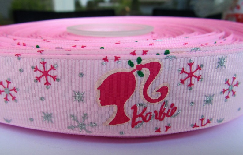 Pu&ograve; includere: Nastro rosa con un motivo a fiocco di neve bianco e una silhouette rosa di Barbie con la parola "Barbie" in lettere argentate scintillanti.