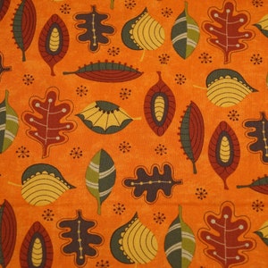 Op de afbeelding: Oranje stof met een herhalend patroon van gestileerde bladeren in tinten bruin, groen en geel. De bladeren zijn in een willekeurig patroon gerangschikt en hebben een retro, mid-century modern esthetiek.