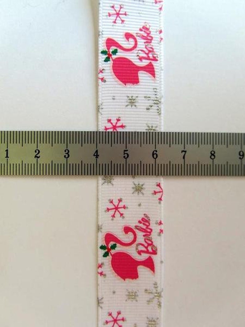 Pu&ograve; includere: Nastro bianco con un motivo di fiocchi di neve argentati glitterati. Il nastro presenta una silhouette rosa di Barbie con la parola "Barbie" stampata in bianco.