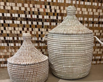 May include: Two woven baskets with lids, featuring a coiled design in alternating white and natural tan fibers. The baskets have a rounded base and a pointed, knob-topped lid. The larger basket includes two small side handles for easy carrying.
