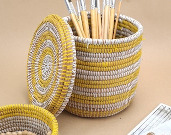 May include: A woven basket with a lid, in yellow and white, holds several paint brushes. A smaller basket with a beaded necklace is in the foreground. The baskets are on a neutral surface.