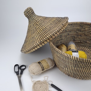 May include: A woven basket with a lid, filled with balls of yarn and a crochet hook. There are also scissors and a crocheted circle on a white surface.