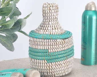 May include: A woven basket with a lid, featuring a white, tan, and turquoise color scheme. The basket is cylindrical with a rounded top and a turquoise band around the middle. Two tubes of product and a green spray bottle are next to the basket.