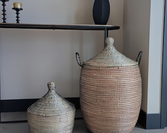 May include: Two woven storage baskets with lids, featuring a natural tan and black pattern. The larger basket is tall and cylindrical with two black side handles, while the smaller basket is shorter and wider with a conical lid and no handles.