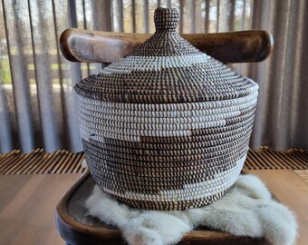 May include: A large, round, woven basket with a lid. The basket is made of natural materials and has a brown and white pattern. The basket is sitting on a wooden chair with a white fur throw.