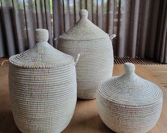 May include: Three white woven baskets with lids. The baskets are made of natural materials and have a traditional design. The largest basket is in the background, and the smallest basket is in the foreground.