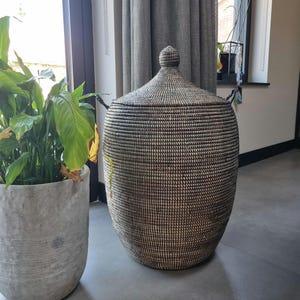 May include: A tall, cylindrical woven basket featuring a dark brown and off-white textured pattern. The basket has a matching conical lid with a small, rounded knob handle at the top and two small side handles for lifting and carrying.