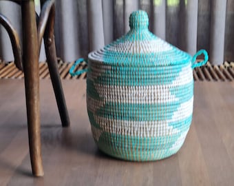 May include: A cylindrical woven basket with a lid and two side handles, featuring a repeating geometric pattern in alternating bands of turquoise and white. The basket has a rounded base and a pointed knob on the center of the lid.