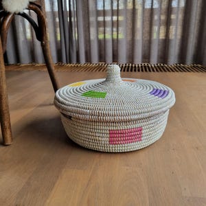 May include: White woven basket with a lid, featuring colorful geometric designs in purple, pink, green, yellow, and orange.