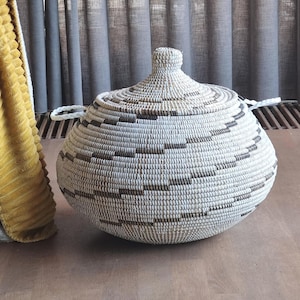May include: A round, woven lidded basket featuring a white base with horizontal bands of dark brown geometric patterns. The basket has a pointed lid with a small knob handle at the top and two white braided handles attached to the sides.
