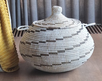 May include: A round, woven lidded basket featuring a white base with horizontal bands of dark brown geometric patterns. The basket has a pointed lid with a small knob handle at the top and two white braided handles attached to the sides.