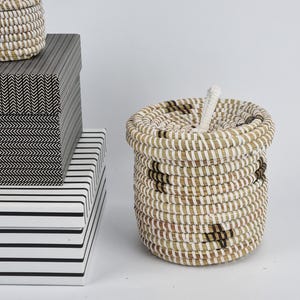 May include: A round woven basket with a lid, alongside a stack of patterned boxes. The basket features a white and brown striped design. The boxes display black and white geometric patterns. The items are presented on a white surface.