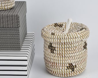 May include: A woven basket with a lid and a stack of decorative boxes. The basket is round with a white and brown striped pattern. The boxes have black and white geometric patterns, including stripes and chevrons. The items are on a white surface.