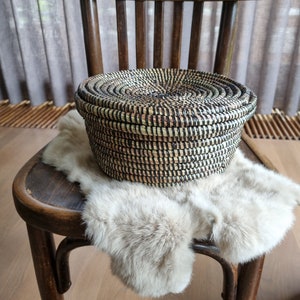 May include: A round, woven basket with a lid, featuring a natural color palette with black accents. The basket is placed on a light-colored fur throw, resting on a vintage wooden chair. The chair has a curved back and legs, adding to the rustic aesthetic.
