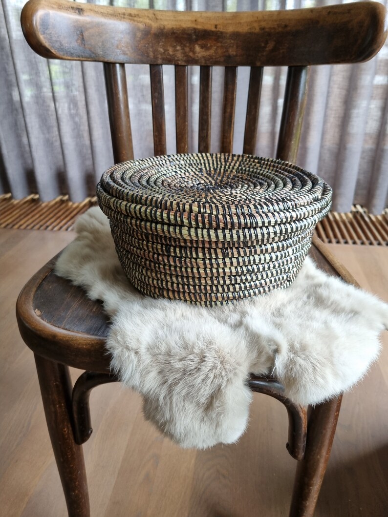 May include: A round, woven basket with a lid, featuring a natural color palette with black accents. The basket is placed on a light-colored fur throw, resting on a vintage wooden chair. The chair has a curved back and legs, adding to the rustic aesthetic.