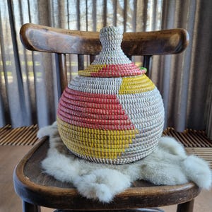 May include: A handwoven basket with a lid, featuring a colorful design of red, yellow, and white stripes. The basket is round and sits on a wooden chair with a fur throw. The basket is approximately 10 inches in diameter.