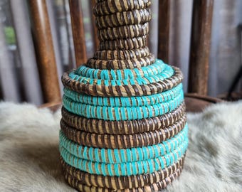 May include: A woven basket with brown and turquoise stripes. The basket is round and has a tall, narrow neck.