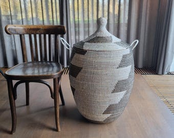 May include: A large, woven storage basket with a lid. The basket is primarily light beige with a geometric pattern in dark brown. It has two small handles and sits on a wooden surface. A wooden chair is to the left.