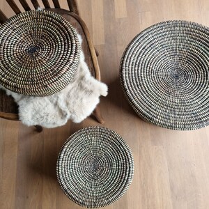 May include: Three round woven baskets in varying sizes. The baskets are made of natural fibers in a spiral pattern with black and green accents. One basket rests on a wooden chair with a faux fur throw, while the others sit on a wooden floor.