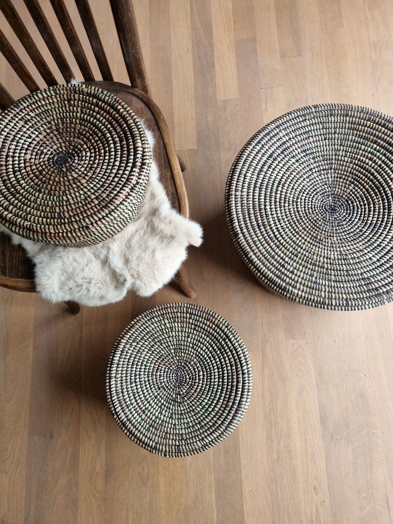 May include: Three round woven baskets in varying sizes. The baskets are made of natural fibers in a spiral pattern with black and green accents. One basket rests on a wooden chair with a faux fur throw, while the others sit on a wooden floor.