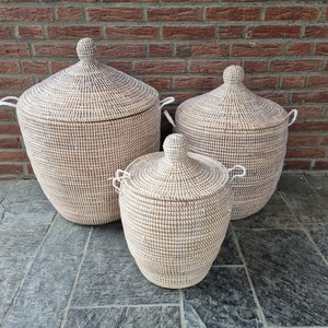 Op de afbeelding: Drie witte en natuurlijke geweven opbergmanden met deksels. De manden zijn rond en hebben een traditioneel ontwerp. De grootste mand is op de achtergrond, en de kleinste mand is op de voorgrond.