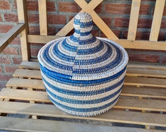 May include: A round, lidded basket with a woven design in alternating bands of blue and natural beige. The basket is sitting on a wooden bench with a brick background. The lid has a knob on top.