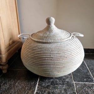 May include: A large, round, woven basket with a matching conical lid and two side handles. The basket features a light, natural off-white color with a textured, coiled construction. It has a wide, bulbous body and a small knob on the lid.