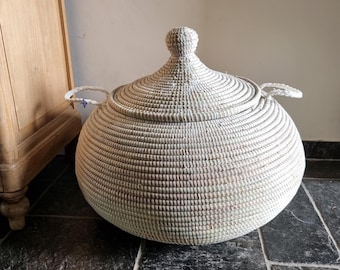 May include: A large, round, woven basket with a matching conical lid and two side handles. The basket features a light, natural off-white color with a textured, coiled construction. It has a wide, bulbous body and a small knob on the lid.