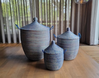 May include: Three woven storage baskets with lids in varying sizes. The baskets are blue and white with blue handles. The lids have a rounded shape with a small knob on top. The baskets are on a wooden surface.