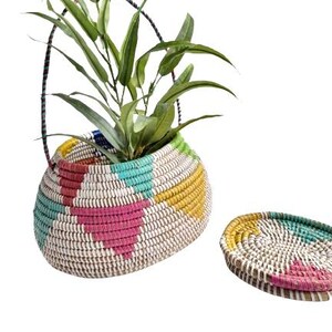 May include: A woven basket planter with a colorful geometric design in pink, teal, yellow, and white, holding a green plant. A matching coaster sits beside it. The basket has a handle.
