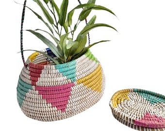 May include: A woven basket planter with a colorful geometric design in pink, teal, yellow, and white, holding a green plant. A matching coaster sits beside it. The basket has a handle.