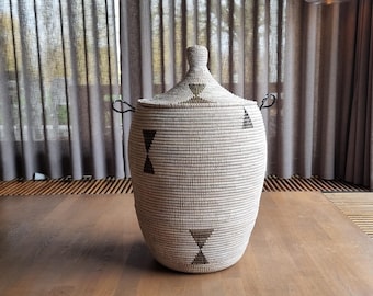 May include: A woven storage basket with a lid, featuring a natural color scheme with dark brown geometric accents. The basket has two black handles and is set on a wooden floor. The background includes curtains and a hanging light fixture.