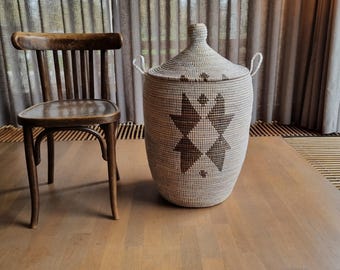 May include: A woven storage basket with a lid, featuring a geometric pattern in brown and cream. The basket has two handles and sits on a wooden floor. A wooden chair is to the left of the basket.
