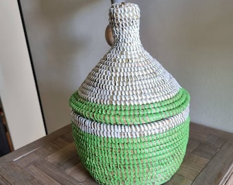 May include: A woven basket with a green and white striped pattern. The basket is round and has a lid. It is sitting on a wooden table.
