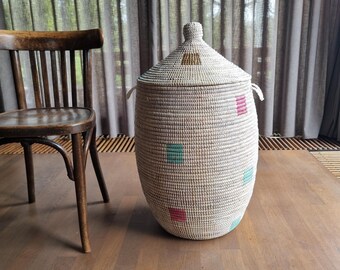 May include: A tall, cylindrical woven basket with a matching conical lid and two side handles. The basket is primarily off-white with small, scattered square accents in muted pink and teal colors, featuring a single brown square on the lid.