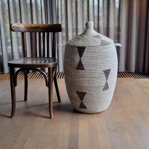 May include: A large, woven basket with a lid, featuring a repeating geometric pattern in brown against a cream background. The basket has two handles and sits on a wooden floor next to a wooden chair. The basket is approximately 61 cm tall.