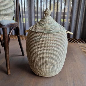 May include: A large, round, natural woven basket with a lid. The basket is made of a light brown material and has a natural, rustic look. The basket is sitting on a wooden floor.