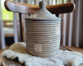 May include: A woven storage basket with a lid, made of natural materials, in a light gray and brown color. The basket is sitting on a furry surface.