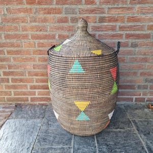 Op de afbeelding: Een grote, geweven mand met een deksel, met een geometrisch patroon van kleurrijke driehoeken in rood, geel, groen en blauw. De mand heeft zwarte handvatten en een bijpassend deksel met een knop. De mand staat tegen een bakstenen muur.