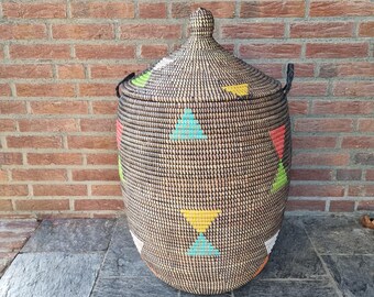 May include: A large, woven basket with a lid, featuring a geometric pattern of colorful triangles in red, yellow, green, and blue. The basket has black handles and a matching lid with a knob. The basket is set against a brick wall.