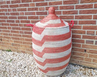 May include: A large, woven storage basket with a lid. The basket is cylindrical with a rounded top and is adorned with horizontal red and white stripes. It has two red handles and a red knob on the lid. The basket is approximately 24 inches tall.