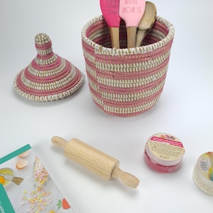 May include: A pink and white woven basket with a lid, holding pink and wooden kitchen utensils. A wooden rolling pin, a jar of pink sprinkles, and a cookbook are also present. The text on the spatula reads "HOME IS WHERE MOM IS."