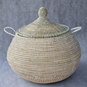 May include: A round, woven basket with a matching conical lid and two white braided handles. The basket is constructed from natural tan fibers interwoven with thin white vertical strips, creating a textured, striped pattern across the surface.