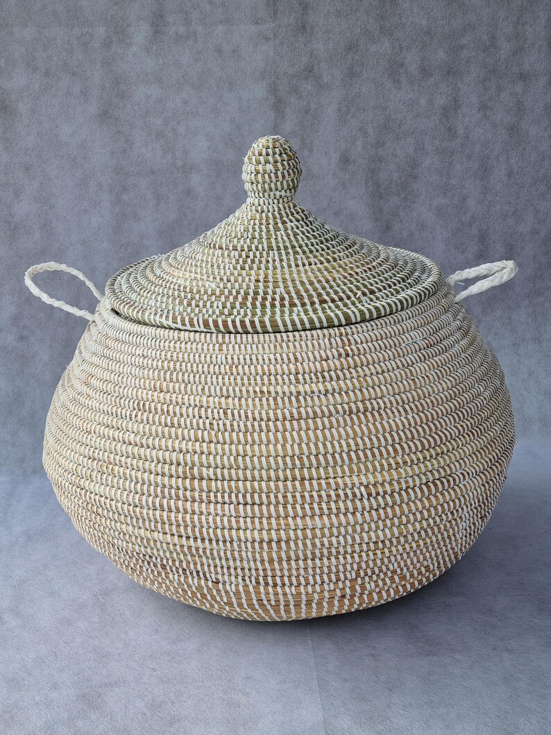 May include: A round, woven basket with a matching conical lid and two white braided handles. The basket is constructed from natural tan fibers interwoven with thin white vertical strips, creating a textured, striped pattern across the surface.