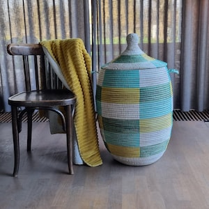 May include: A woven storage basket with a lid, featuring a geometric pattern in shades of green, blue, yellow, and gray. A yellow blanket is draped over a wooden chair. The basket has two handles and a rounded top.