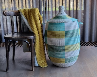 May include: A woven storage basket with a lid, featuring a geometric pattern in shades of green, blue, yellow, and gray. A yellow blanket is draped over a wooden chair. The basket has two handles and a rounded top.