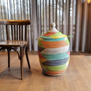 Op de afbeelding: Een grote, geweven opbergmand met een deksel. De mand is cilindrisch en heeft een levendig patroon van horizontale strepen in verschillende kleuren, waaronder oranje, groen, blauw en geel. Hij heeft twee kleine handvatten en staat op een houten vloer.