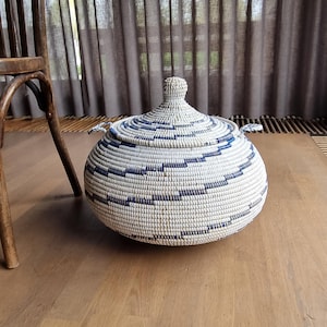 May include: A round, lidded basket with a woven design in white and navy blue. The basket sits on a wooden floor, with a wooden chair and a curtain in the background. The lid has a small knob on top and two small handles.