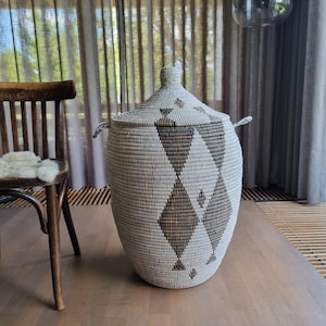 May include: A large, woven storage basket with a lid. The basket is primarily off-white with a repeating pattern of brown diamond shapes. It has two small handles on the sides and a handle on the lid. The basket is sitting on a wooden floor.